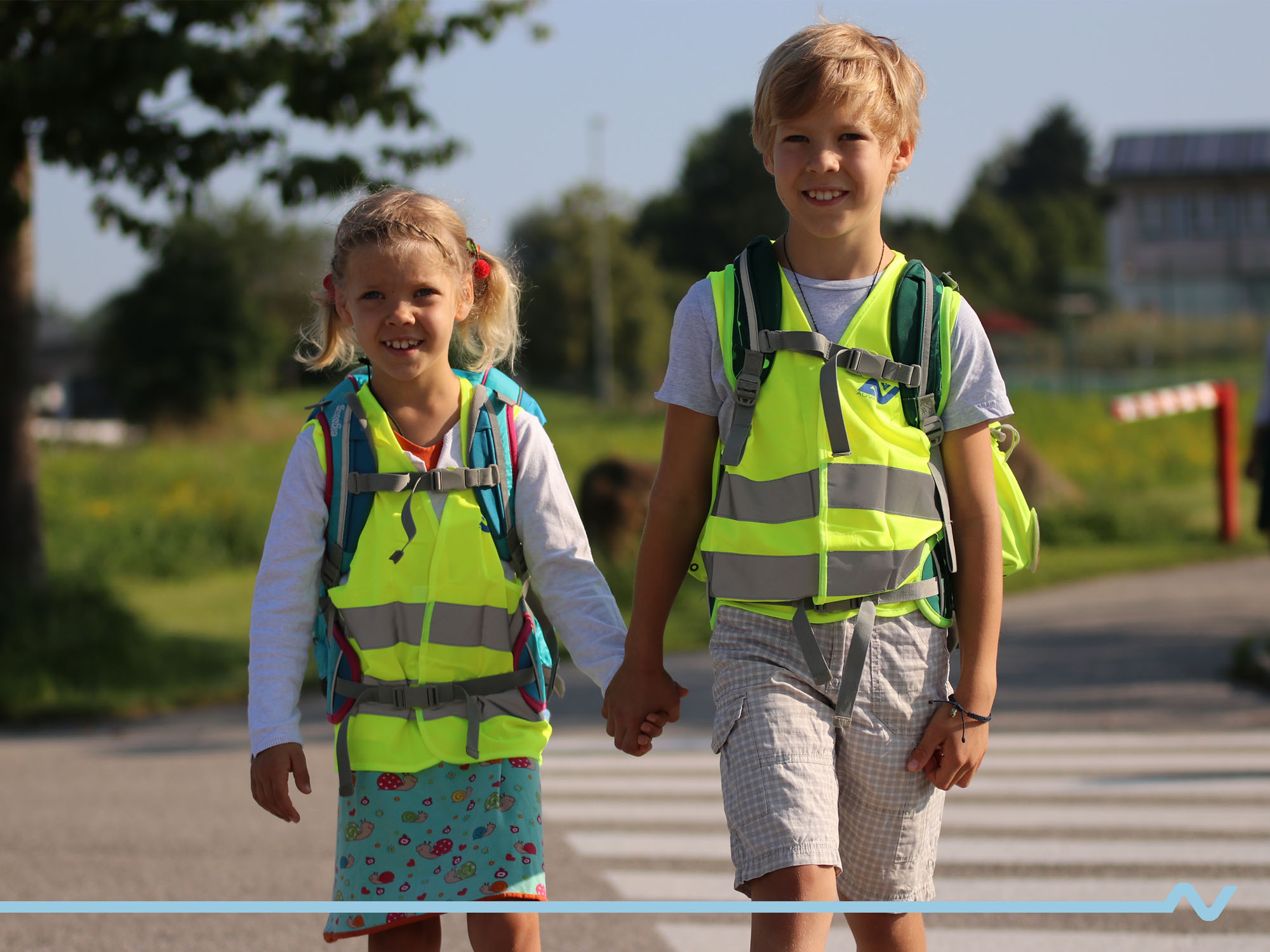 Ein Mädchen und ein Bub gehen mit Warnwesten und Schultaschen bekleidet Hand in Hand zur Schule