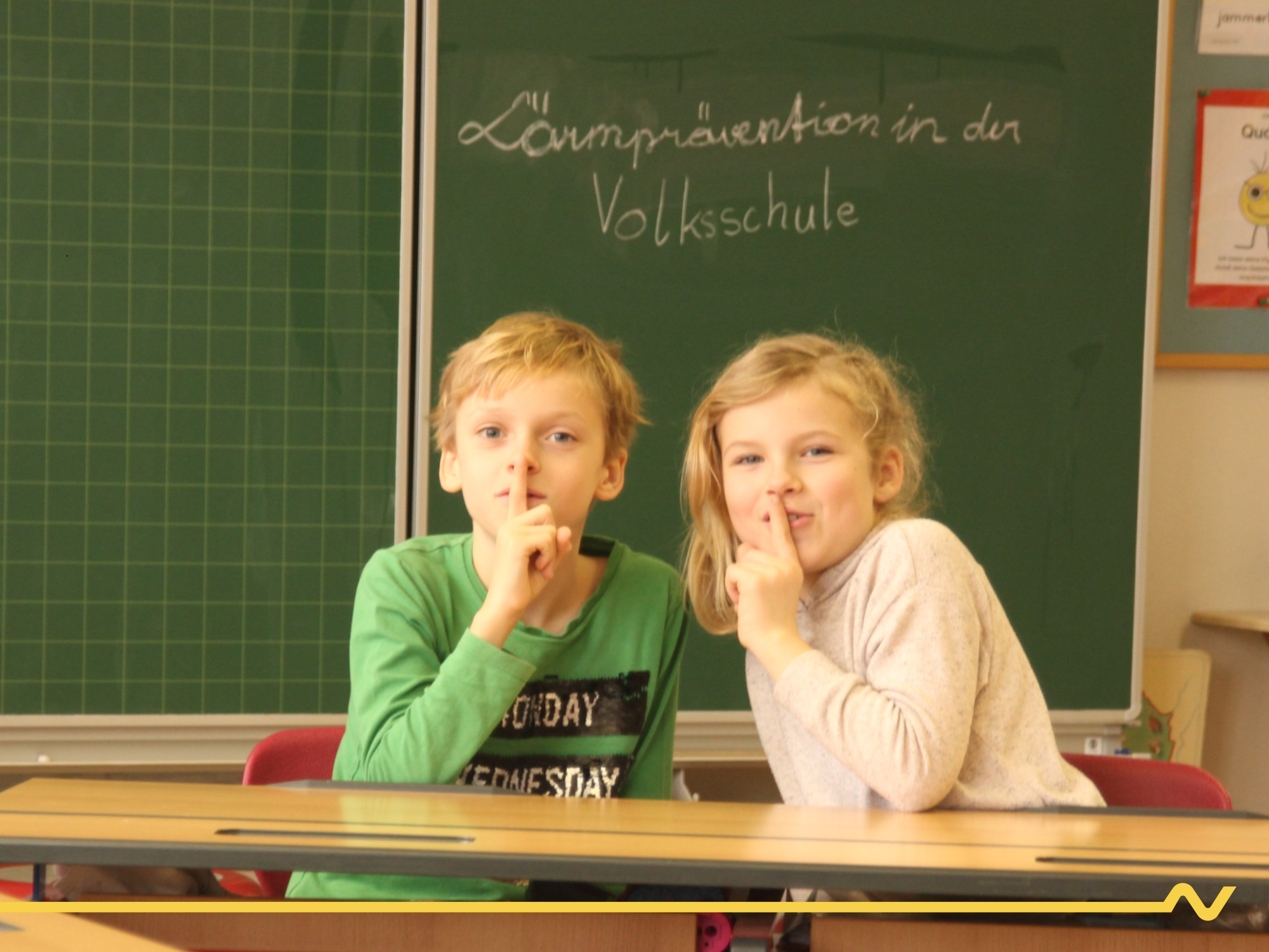 Zwei Volksschulkinder sitzen vor der Tafel im Klassenzimmer und deuten mit dem Zeigefinger die "Leise"-Geste. Auf der Tafel im Hintergrund steht "Lärmprävention in der Volksschule"